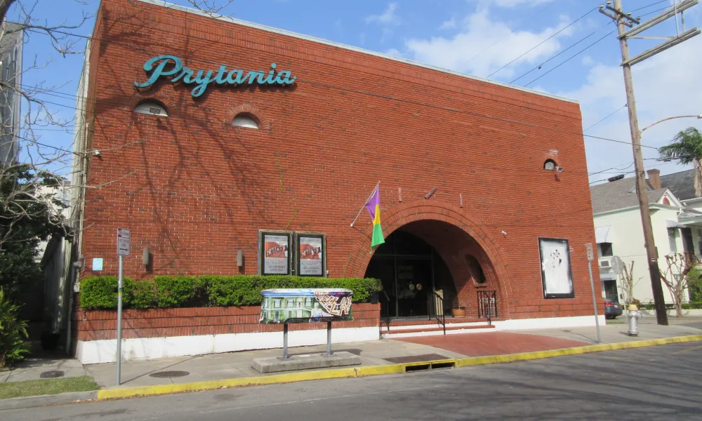 Red brick building with arched entrance and Prytania sign under blue sky, surrounded by greenery and street elements.