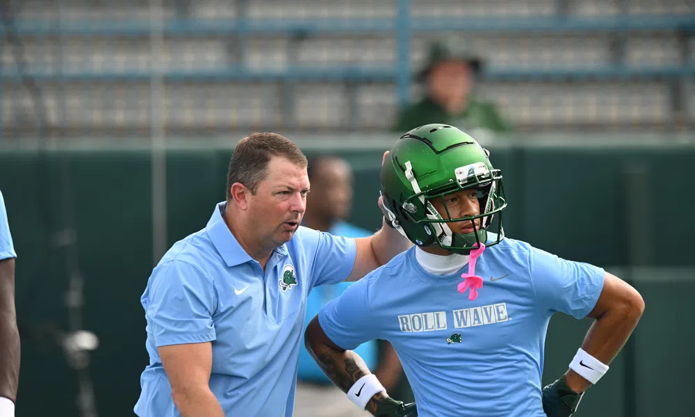 Football coach instructing player wearing green helmet and blue Roll Wave shirt during practice session