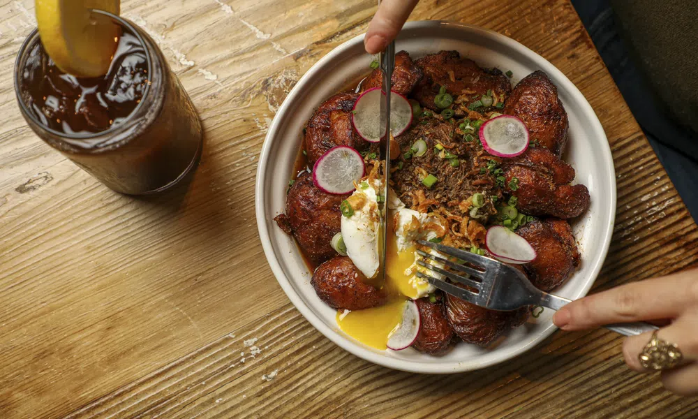 Bowl of fried plantains with poached egg, shredded meat, and radish slices on a wooden table with iced tea.