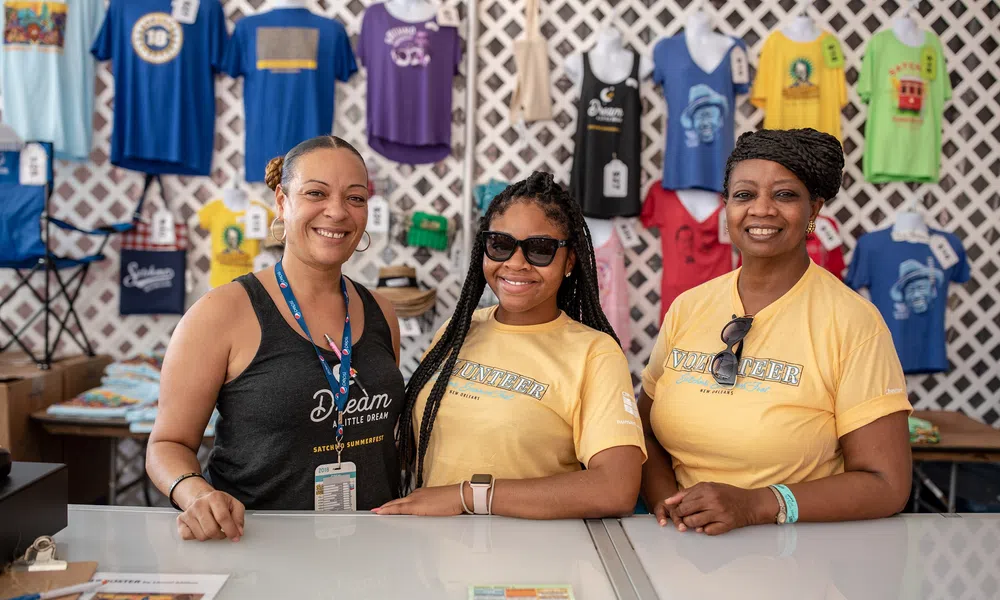 Three smiling women standing behind a counter with colorful t-shirts hanging on the wall behind them