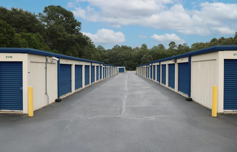 Outdoor self-storage units with blue roll-up doors on both sides and a paved central driveway under a partly cloudy sky.