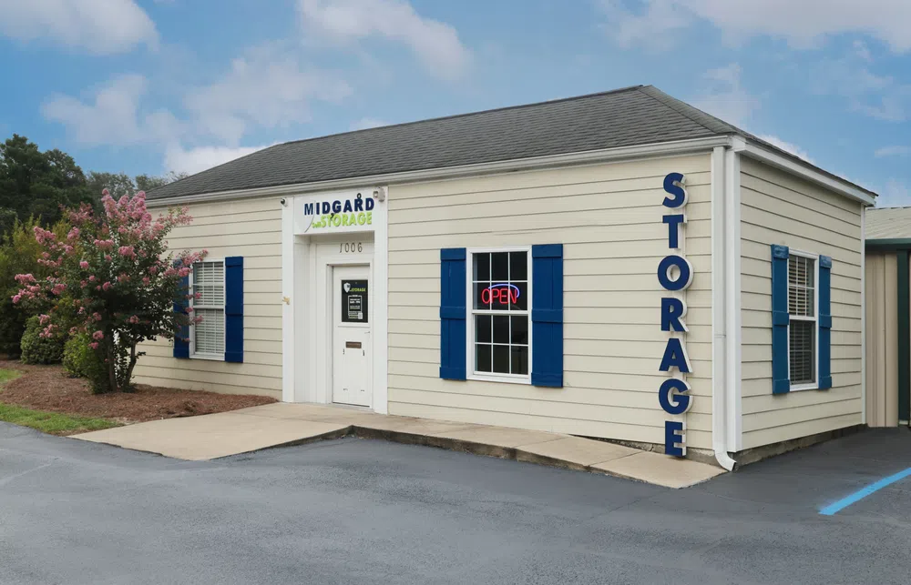 Beige Midgard Storage building with blue shutters, open sign, and pink flowering tree in front under a blue sky