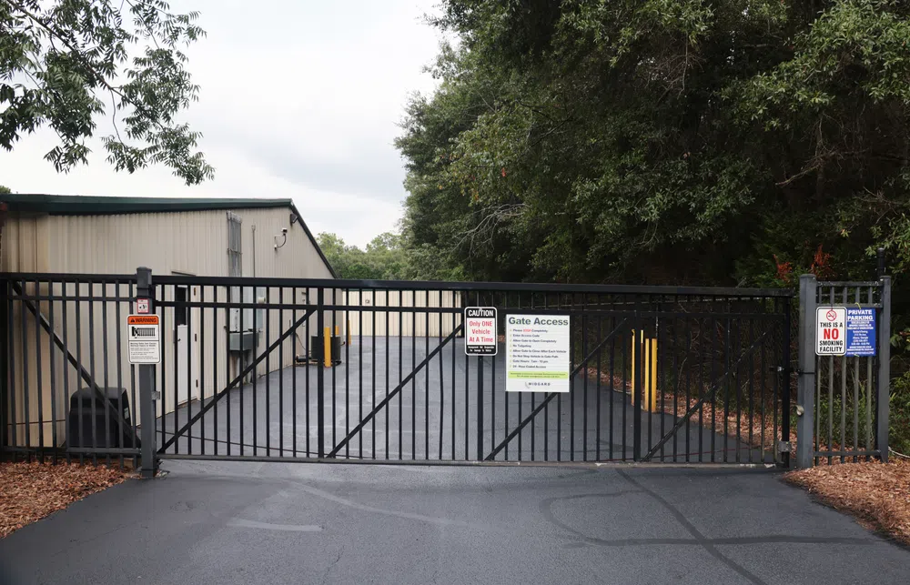 Black metal security gate blocking driveway access to storage facility with warning and gate access signs.