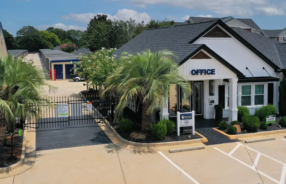Storage facility office building with gated access, palm trees, and parking spaces under a blue sky.