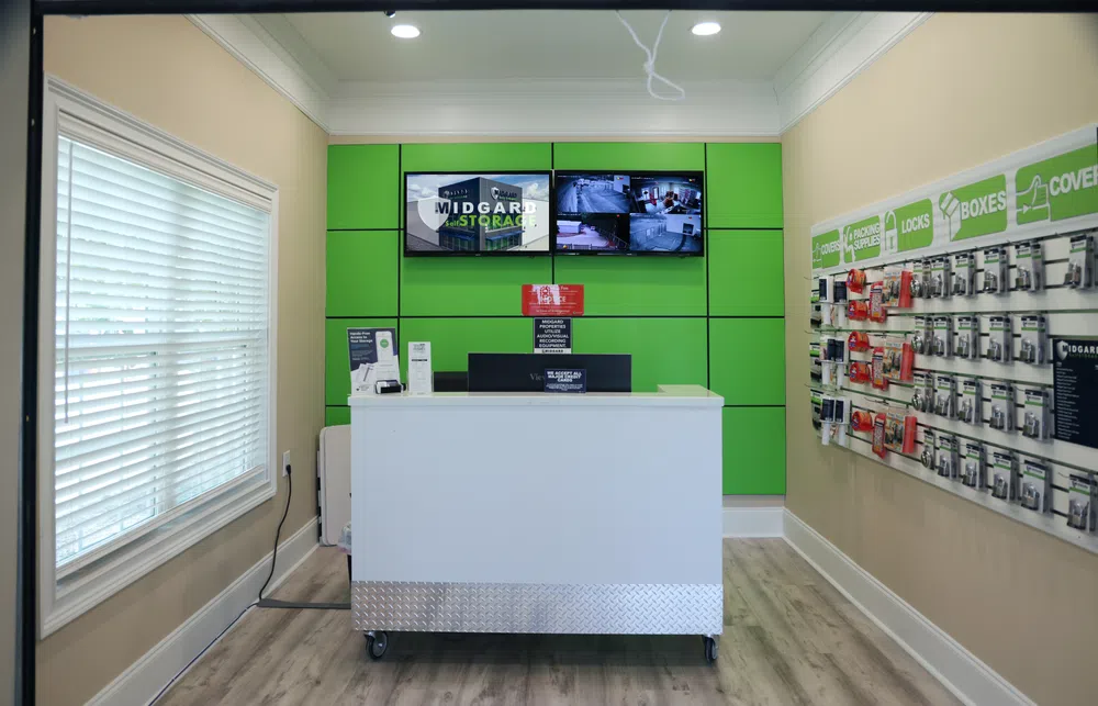 Modern storage facility office with green wall panels, white counter, and security monitors displaying surveillance feeds.