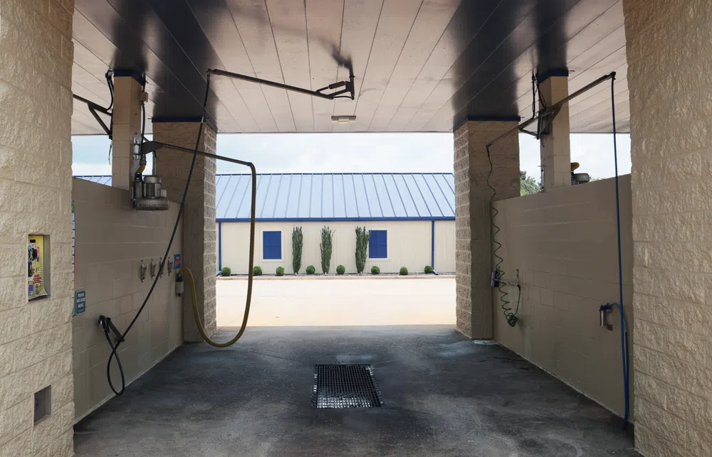 Empty self-service car wash bay with hoses and drains under a covered roof near a building with a blue roof.