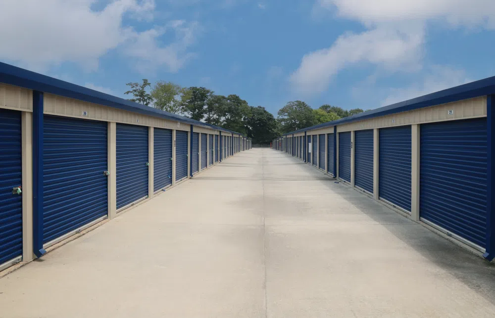 Long central pathway flanked by rows of blue storage unit doors under a partly cloudy sky.