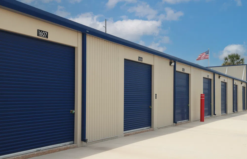 Row of blue roll-up doors on beige storage units under a clear sky with an American flag in the background