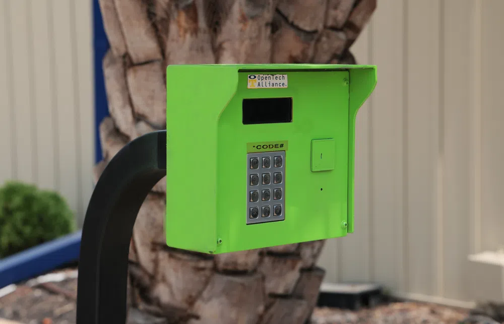 Green keypad access control box on black stand with tree and building background outdoors