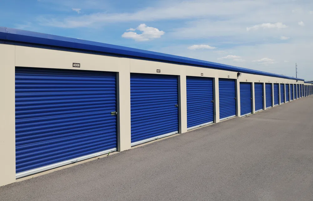 Row of storage units with blue roll-up doors under a clear sky on an asphalt driveway.