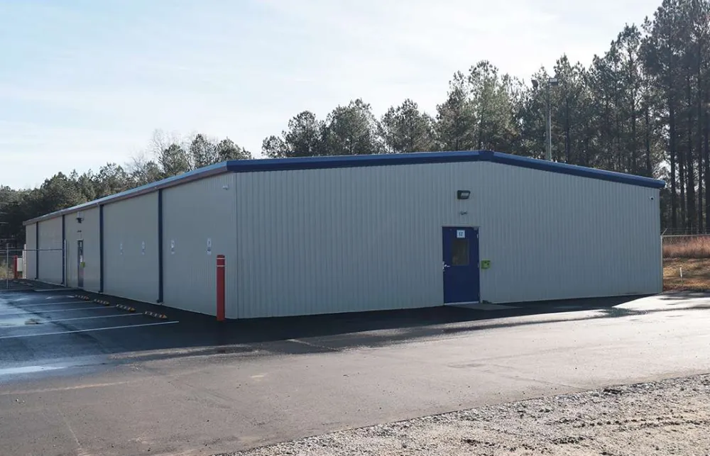 Single-story industrial metal building with blue trim beside a paved parking lot under clear sky near trees.
