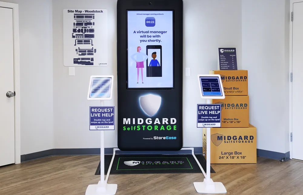 Self-storage kiosk with virtual manager screen, request live help tablets, and stacked Midgard storage boxes in office lobby.