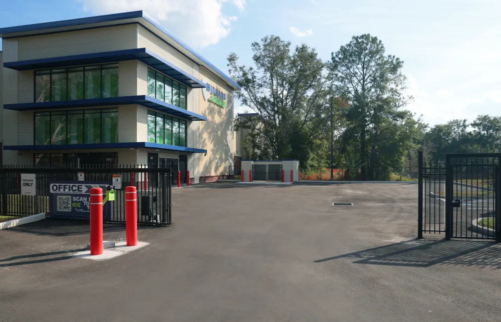 Modern storage facility with gated entrance, office building, and surrounding trees under blue sky.