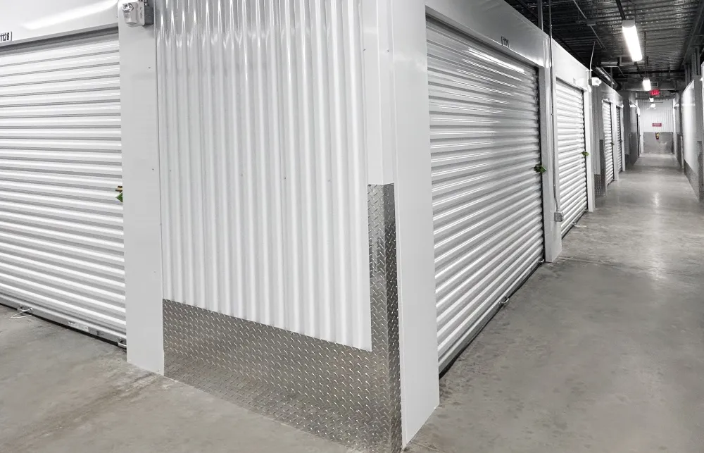 Indoor storage facility corridor with white roll-up metal doors and concrete floor under fluorescent lighting