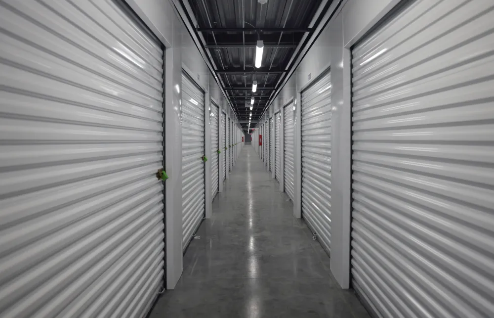 Long corridor of indoor storage units with closed white roll-up doors and polished concrete floor.