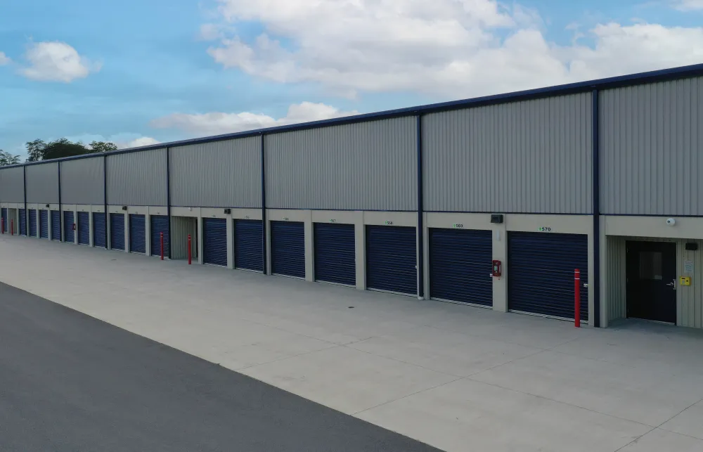 Row of closed blue storage unit doors on a concrete drive at a self-storage facility under a cloudy sky.