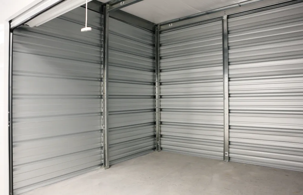 Empty storage unit with metal walls and a concrete floor, showing a partially open roll-up door and pull cord.