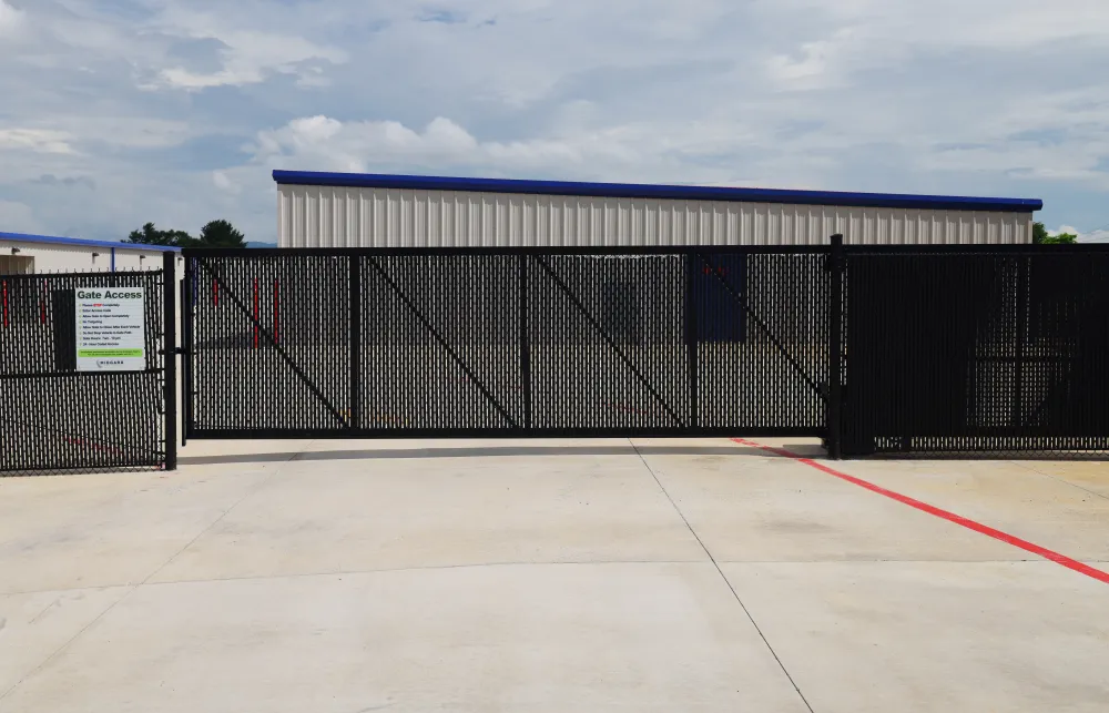 Black metal security gate with access instructions in front of a storage facility building under a cloudy sky.