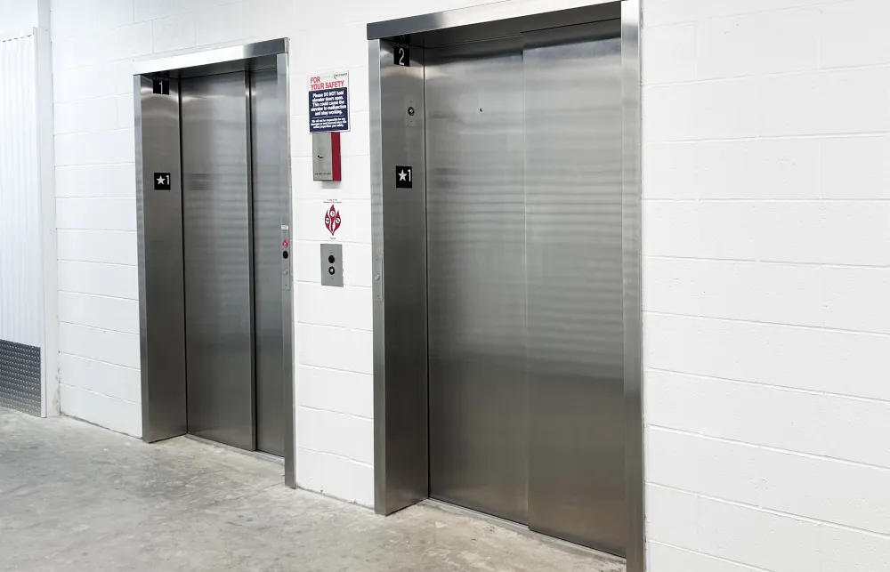 Two closed stainless steel elevator doors in a white indoor hallway with safety signs and buttons.
