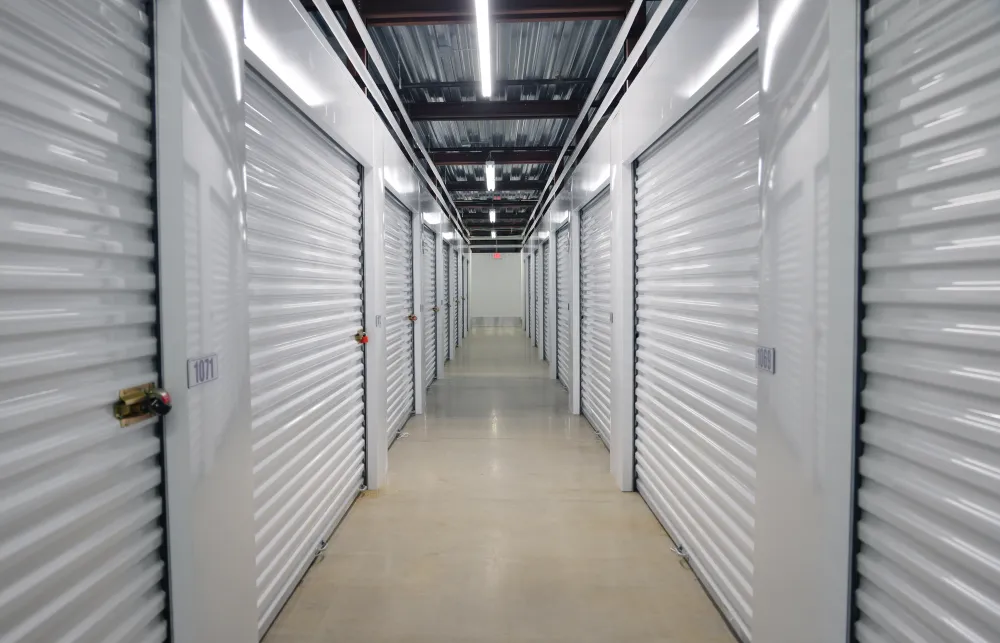 Long corridor with closed white metal storage unit doors and bright overhead lighting in a clean storage facility.