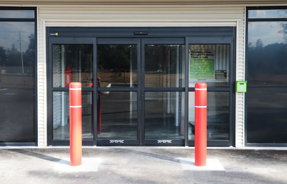 Automatic sliding glass doors with red bollards and a keypad access system outside a storage facility.