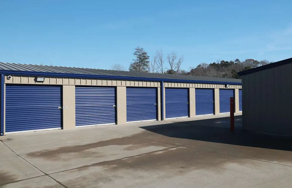 Outdoor row of storage unit garages with blue roll-up doors under a clear sky on concrete pavement.
