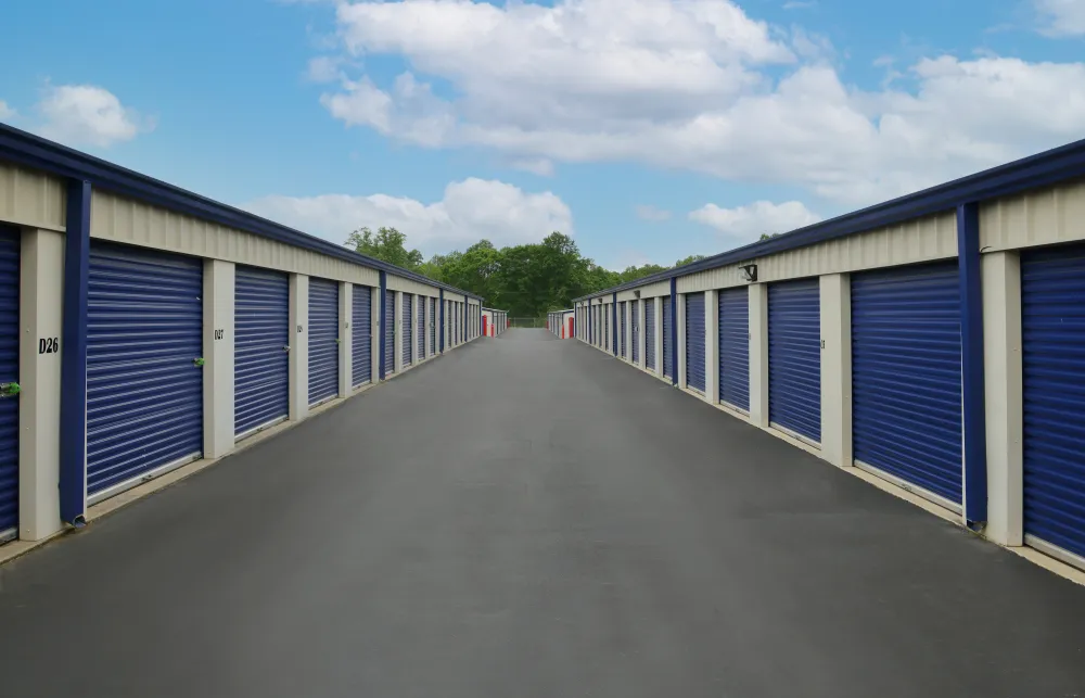 Outdoor self storage units with blue roll-up doors lining a paved driveway under a partly cloudy sky.