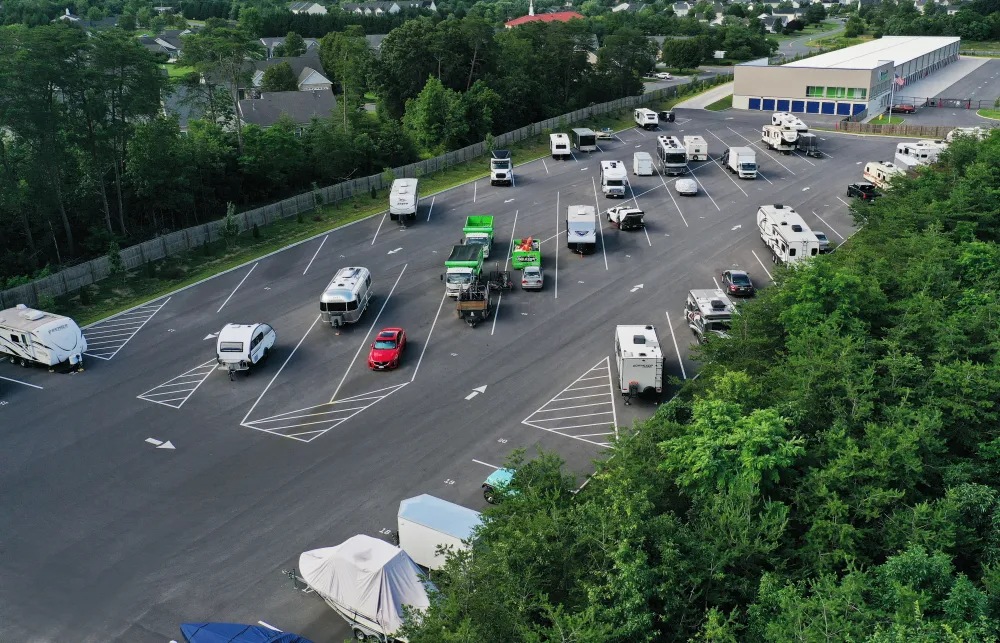 Large parking lot with various RVs, trailers, and vehicles surrounded by trees and nearby buildings on a clear day