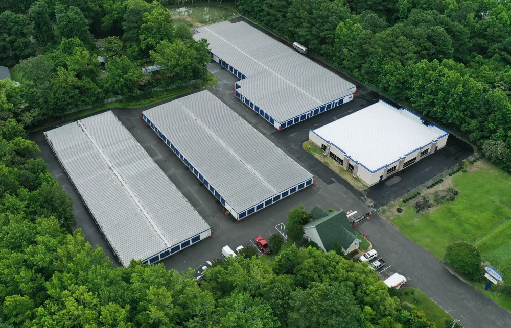 Aerial view of a storage facility with multiple large buildings surrounded by dense green trees and parked vehicles.