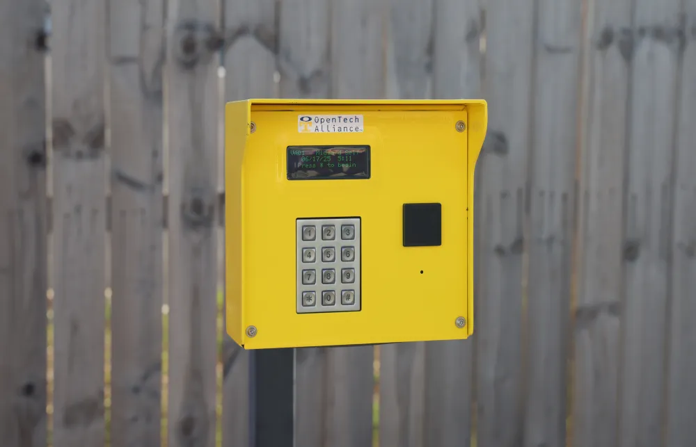 Yellow access control keypad mounted on a post in front of a wooden fence with OpenTech Alliance label.