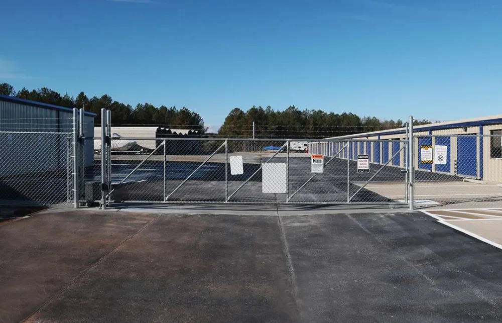 Chain link security gate closed at the entrance of a large outdoor storage facility with storage units and clear blue sky.