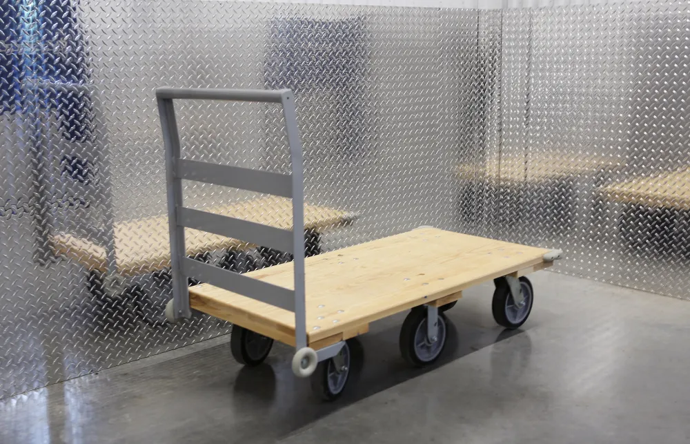 Flat wooden platform cart with metal handle and four wheels on a reflective steel floor and wall