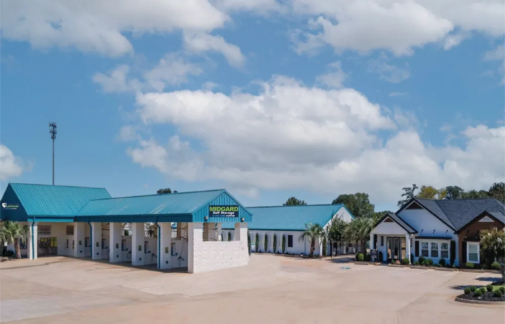 Midgard Self Storage facility with blue roofs, multiple buildings, palm trees, and clear sky with clouds.