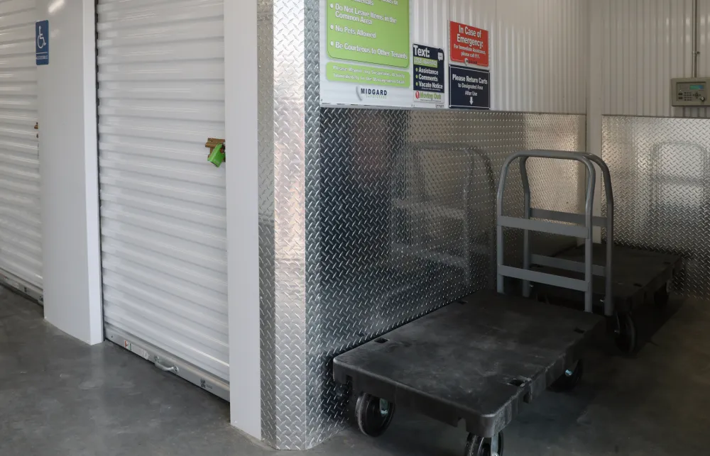 Storage unit hallway with closed white roll-up door, caution signs, and two black flatbed carts on concrete floor.