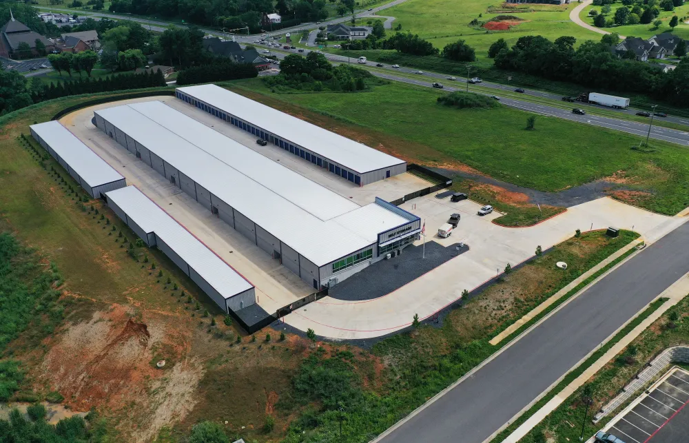 Aerial view of a large self storage facility with multiple white-roofed buildings near a highway and green fields.