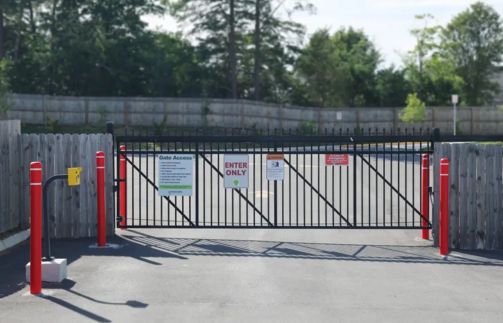 Closed black metal gate with signs and red bollards on paved entrance with wooden fence and trees in background