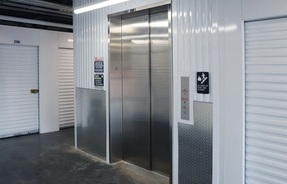 Stainless steel elevator doors in a clean storage facility hallway with white roll-up storage unit doors.