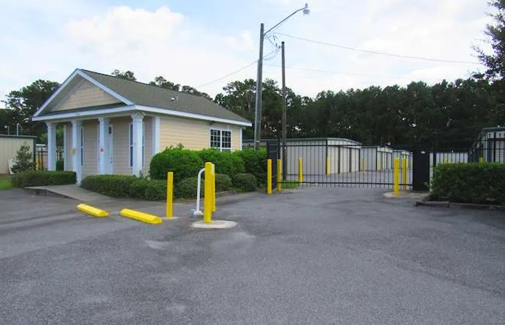 Gated storage facility entrance with small yellow building and multiple storage units behind black security gate.