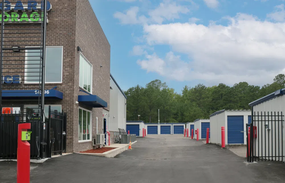 Outdoor storage facility with blue roll-up doors, gated entrance, and clear sky with trees in the background
