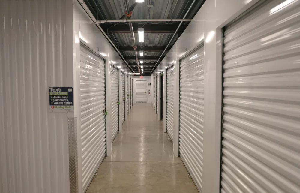 Indoor hallway with rows of white metal storage unit doors in a well-lit storage facility.