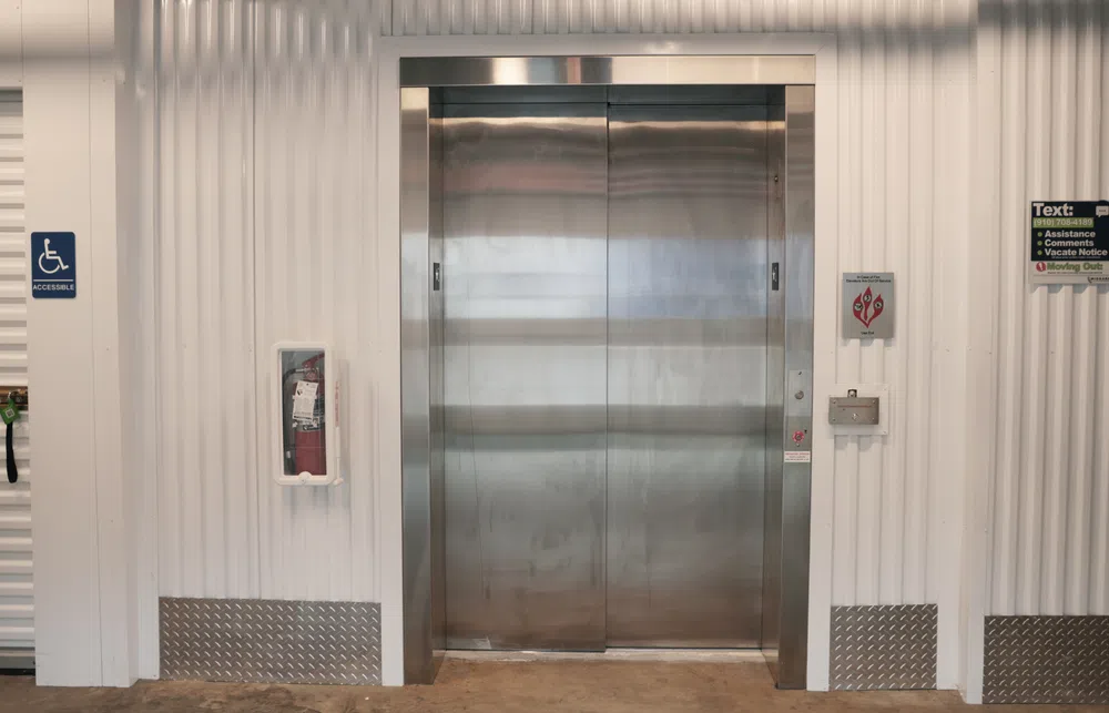 Metal elevator doors in a white industrial corridor with accessible signage and fire safety equipment nearby.