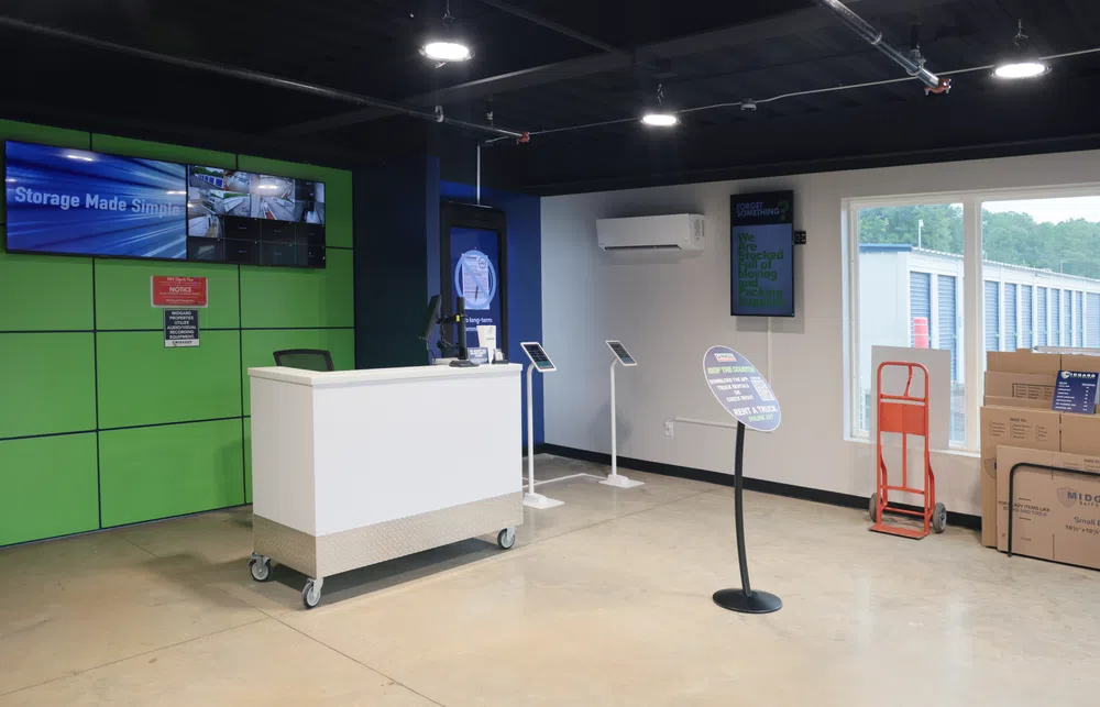 Modern self-storage office with white counter, video screens, storage boxes, and dolly near large window.