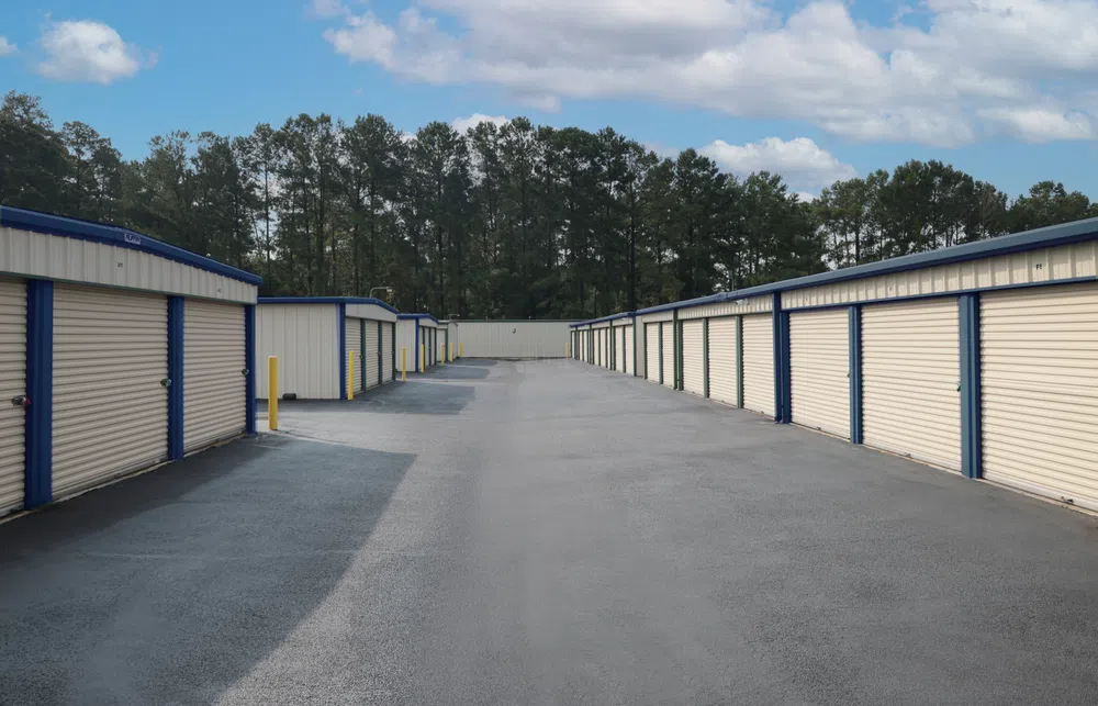 Empty outdoor storage facility with rows of closed beige and blue storage unit doors under a partly cloudy sky.