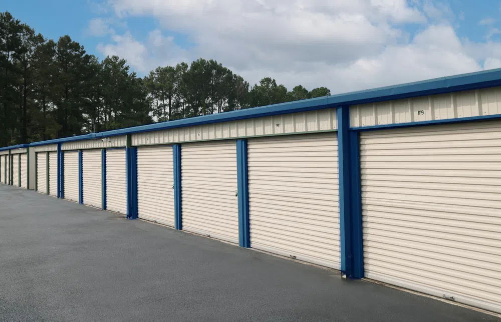 Row of beige and blue storage unit doors under a partly cloudy sky with trees in the background.