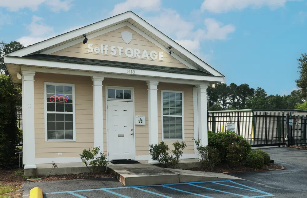 Small beige self-storage office building with white trim and an open sign, adjacent to gated storage units.