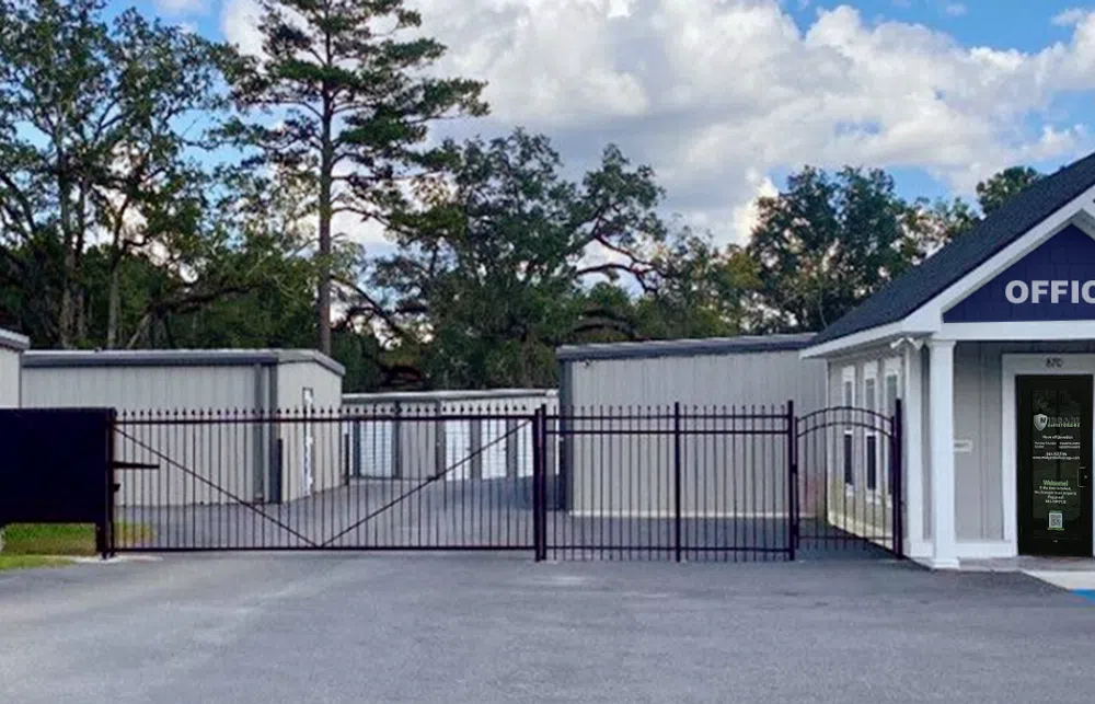 Gated storage facility with gray units, black iron gate, and a small office building under a partly cloudy sky.