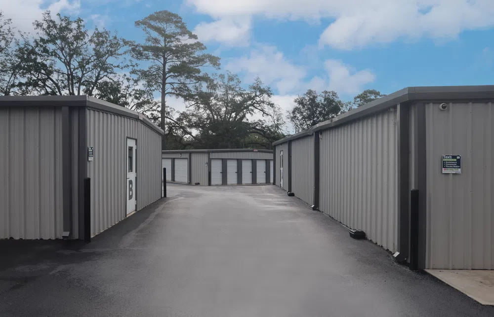 Outdoor view of a storage facility with metal storage units along a paved driveway under a partly cloudy sky.