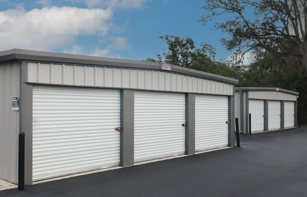 Gray metal storage units with white roll-up doors along a paved driveway under a blue sky with clouds.