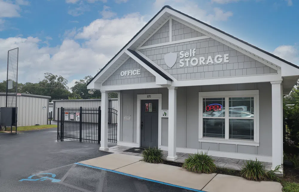 Gray self storage office building with white trim, columns, and an open sign in the window under a blue sky.