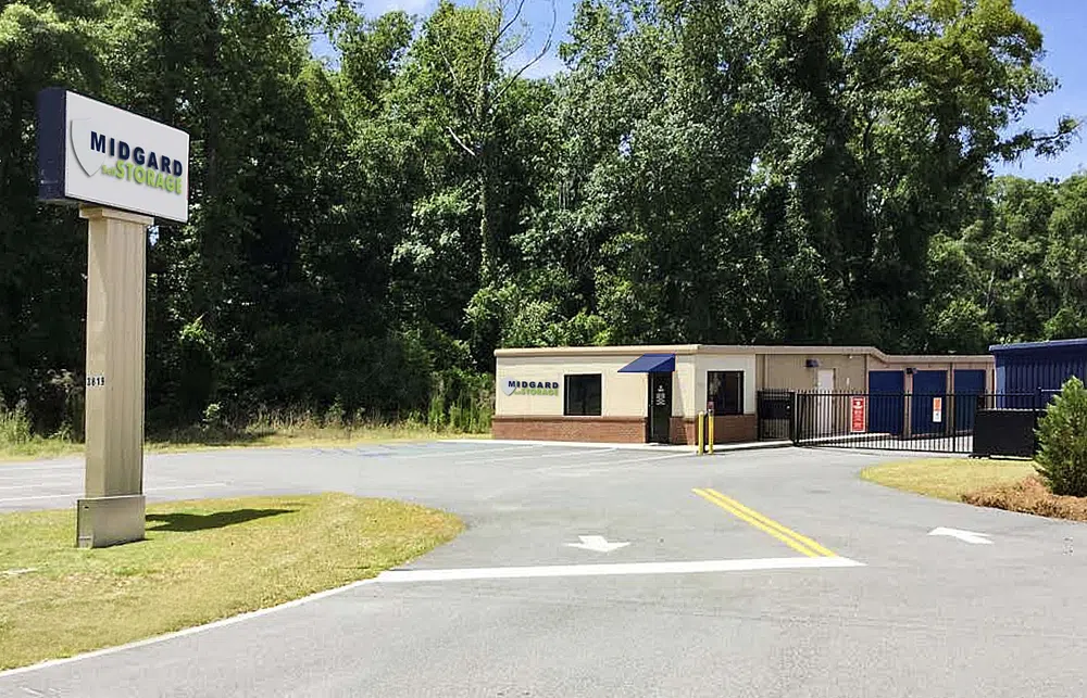 Midgard Mini Storage facility with sign, office building, gated entrance, and surrounding trees under blue sky